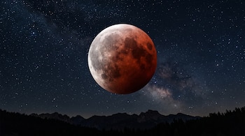 Luna llena rojiza cubierta parcialmente por una sombra, ubicada sobre un cielo estrellado y la silueta de montañas.