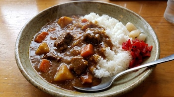Un plato de Karē Raisu con curry espeso de carne y verduras, arroz blanco al vapor, y dos tipos de encurtidos, sobre una mesa de madera.