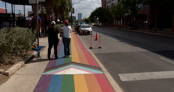 Dos personas caminan sobre una acera pintada con los colores del arcoíris y la bandera del orgullo, junto a una calle con coches estacionados y conos de tráfico