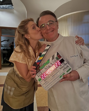 Una mujer le da un beso en la mejilla a un hombre con anteojos y bata médica que sostiene una claqueta de cine