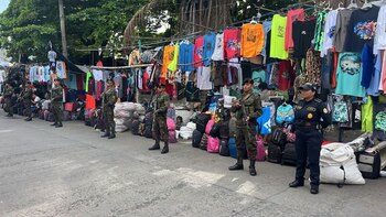 Vista de calle con varios soldados uniformados con rifles y una policía de pie frente a puestos de ropa y bolsos colgados, con un niño sentado entre los artículos