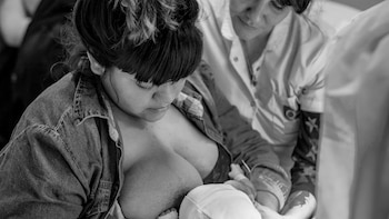 Imagen en blanco y negro de una madre con camisa de mezclilla amamantando a su recién nacido con gorro blanco. Una persona con uniforme claro asiste en segundo plano