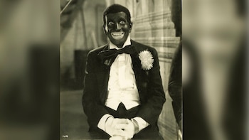 Hombre con el rostro pintado de negro, amplia sonrisa, esmoquin, pajarita, una flor en la solapa y guantes blancos en foto en blanco y negro