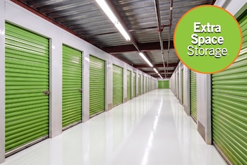 Un pasillo de depósitos con hileras de puertas metálicas verdes, suelo blanco, techo de metal y luces. Un logo circular verde dice Extra Space Storage