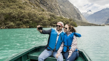 Una pareja sonríe en un bote sobre un lago turquesa. El hombre toma una selfi con su teléfono. Visten chalecos salvavidas azules con montañas al fondo
