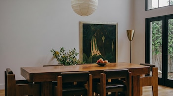 Un comedor luminoso con una gran mesa de madera y sillas de cuero, una lámpara de techo de papel y una pintura de un bosque en la pared. Un cuenco de frutas y un florero decoran la mesa. Una puerta de cristal ofrece vista a la vegetación.