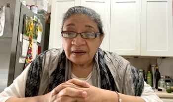 Retrato de Odalys González Silvera, una mujer de 61 años con gafas, camiseta blanca y bufanda estampada, mirando al frente con las manos entrelazadas, con un fondo de cocina