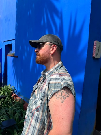 Hombre de perfil, barba, gorra, gafas de sol, aro. Camisa a cuadros sin mangas, tatuaje de dos rostros besándose en el brazo. Pared azul, plantas, cartel de baño de mujeres