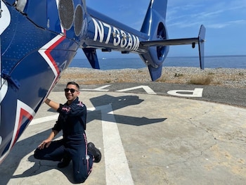 Un hombre con gafas de sol y uniforme azul se arrodilla junto a un helicóptero azul en un helipuerto frente al mar