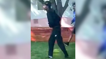 Un joven con gorra, sudadera y pantalón oscuro camina sobre césped. Su perfil lateral es visible, con un fondo borroso de árboles y estructuras