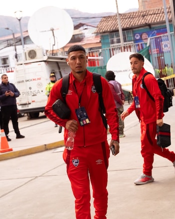 Dos futbolistas de Cienciano con uniformes deportivos rojos y mochilas. Uno sostiene una botella de agua. Detrás, una van de prensa con antenas