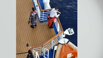 Vista aérea de la cubierta de un barco con tres personas. Una lleva chaleco salvavidas, otra se inclina sobre la barandilla, y una tercera levanta el brazo. Mar azul y bote salvavidas