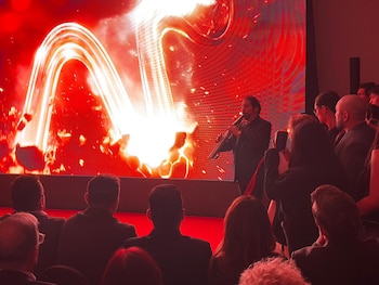 Un músico toca un instrumento de viento oscuro frente a una pantalla grande con intensos gráficos abstractos rojos y blancos; la audiencia se ve de espaldas