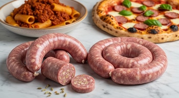 Salsiccia italiana cruda, algunas enrolladas y una rebanada con semillas de hinojo, sobre un mostrador de mármol. Al fondo, pasta con ragú y una pizza con salsiccia.