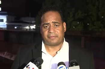 Un hombre con saco negro y camisa blanca habla frente a tres micrófonos con logotipos. El fondo es oscuro y difuso con algunas luces
