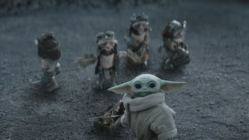 Grogu, un pequeño ser verde con orejas grandes, tira de un carrito sobre tierra oscura, con cuatro criaturas pequeñas con vestimentas de corteza detrás