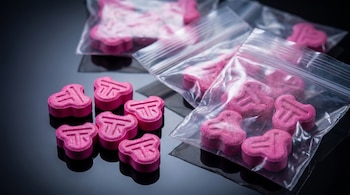 Primer plano de pastillas rosa de Tusi con un símbolo, algunas sobre una superficie reflectante oscura y otras en bolsas de plástico resellables.