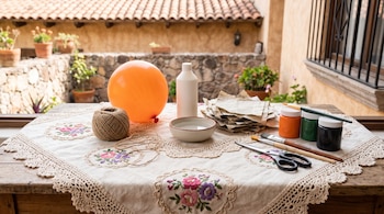 Un globo naranja, un ovillo de cuerda, pegamento, agua, trozos de papel, tarros de pintura, pinceles y tijeras sobre un mantel.