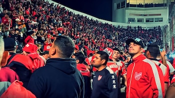 Gran multitud de aficionados con camisetas rojas y blancas en un estadio deportivo nocturno, algunos tocando instrumentos de viento y otros mirando hacia arriba