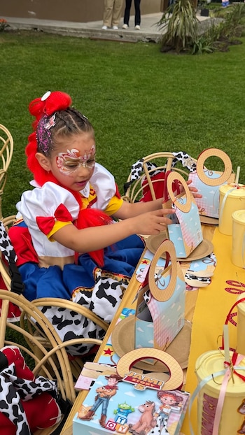 Una niña disfrazada como el personaje Jessie de Toy Story manipula una bolsa de recuerdos sobre una mesa decorada en un jardín