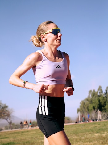 Mujer rubia de perfil con gafas de sol amarillas, top lavanda y pantalón corto negro, corre en pista de tierra con césped y árboles