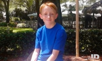 Retrato de un joven de piel clara y cabello rojo, vestido con una camiseta azul. Está sentado al aire libre frente a un fondo borroso de árboles y arbustos verdes