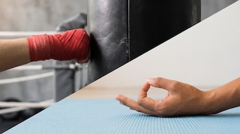 Mano con venda deportiva roja impacta bolsa de boxeo negra en gimnasio; mano en posición de mudra sobre colchoneta azul claro.