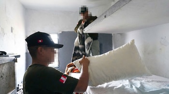 Dos uniformados con insignias peruanas revisan una almohada y mantas sobre literas de concreto en el interior de una celda.