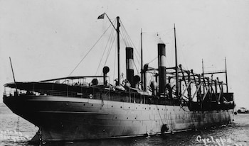 Fotografía en blanco y negro de un gran barco militar con chimeneas, mástiles y la bandera de Estados Unidos navegando en el agua