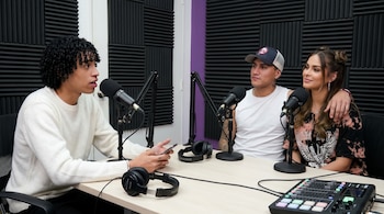 Três pessoas sentadas em uma mesa com microfones e fones de ouvido em um estúdio de podcast, com painéis acústicos na parede posterior. Um deles está segurando um celular.