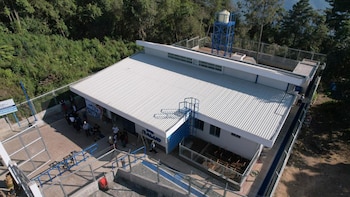 Vista aérea de un moderno centro de salud con techo blanco corrugado y detalles azules, un tanque de agua elevado y personas en la entrada, rodeado de árboles