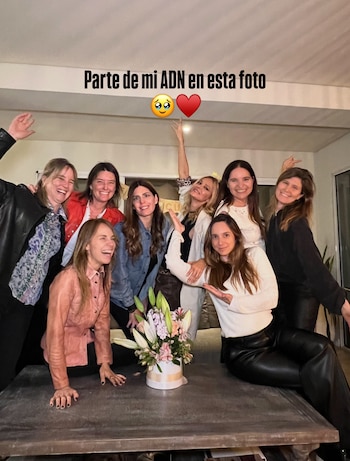 Ocho mujeres posan sonrientes en una sala detrás de una mesa de centro que sostiene un florero blanco con lirios