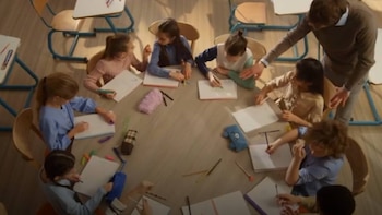 Vista aérea de un maestro supervisando a varios niños sentados alrededor de una mesa redonda, todos escribiendo o dibujando en libretas con materiales escolares