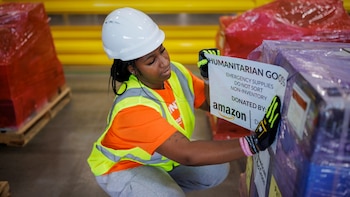 Mujer con casco blanco, chaleco de seguridad amarillo y naranja, y guantes, aplica etiqueta a un palé de carga. La etiqueta indica "Humanitarian Goods" y "Amazon"