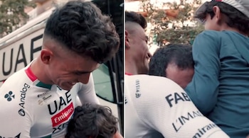 Un ciclista con uniforme blanco del UAE Team Emirates abraza a un adulto con cabello rizado oscuro y a un niño con camiseta azul y gorra blanca.