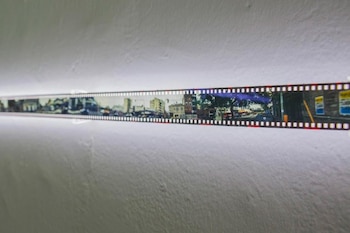 Primer plano de una tira de película horizontal iluminada sobre una pared blanca, mostrando múltiples fotogramas con escenas de calles y edificios