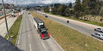 Una estación de peaje con un letrero azul de tarifas, señales de tránsito, varios automóviles en la vía y un ciclista en primer plano