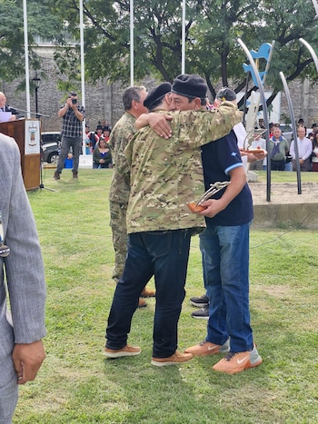 Dos hombres se abrazan en un evento al aire libre sobre césped. Uno viste uniforme camuflado y boina negra, el otro polo azul y jeans, sosteniendo un trofeo plateado