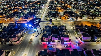 Vista aérea nocturna de un dron policial con luces encendidas que vuela sobre calles iluminadas y tres patrullas aparcadas en un cruce.