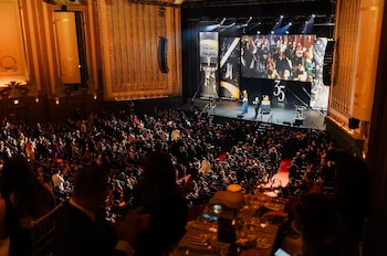 Vista elevada de un auditorio opulento, lleno de asistentes a un evento formal, con un escenario brillante mostrando presentadores, pantallas y decoraciones
