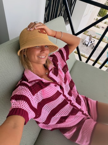 Renata Mónaco sonríe sentada en un sofá, con un sombrero de paja y un conjunto tejido a crochet rosa y bordo, frente a un balcón y vegetación