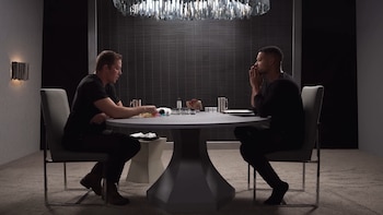 Dos hombres sentados frente a una mesa redonda en un cuarto oscuro y moderno. Uno mira objetos en la mesa; el otro tiene las manos juntas bajo la barbilla