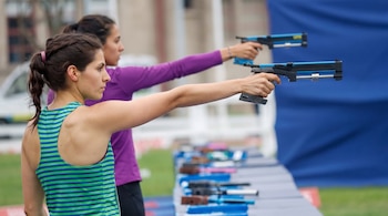 Dos mujeres apuntan de perfil con pistolas láser azules sobre una mesa con equipos de tiro en un campo al aire libre.