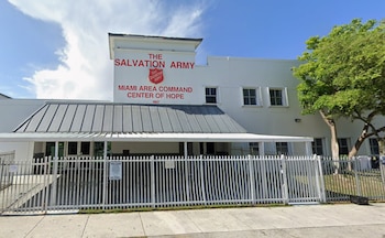 Fachada de un edificio blanco del Ejército de Salvación con letrero rojo, reja metálica blanca y un árbol al lado