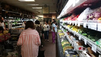 Interior de un supermercado con clientes comprando. Estantes refrigerados llenos de pimientos, limones, calabacines y vegetales verdes. Carritos de compra