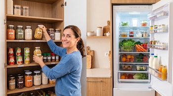 Una mujer sostiene un frasco frente a estantes con legumbres y verduras junto a un refrigerador abierto repleto de alimentos.