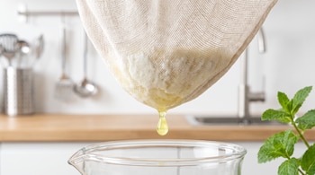 Bolsa de tela suspendida que contiene masa blanca de kéfir y deja caer una gota de suero en un recipiente de vidrio.