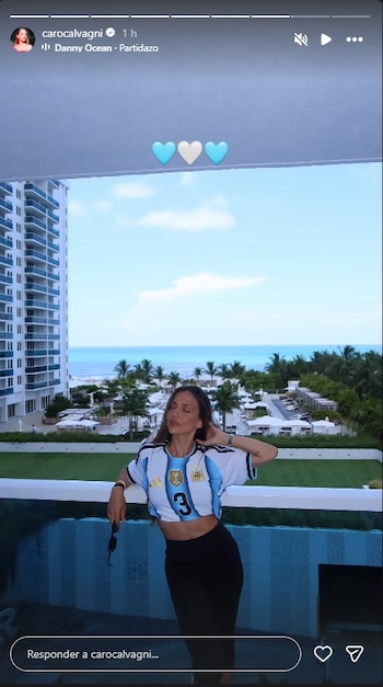 Mujer en balcón con camiseta de la selección argentina y falda negra, ante un edificio, palmeras, playa y mar bajo cielo celeste