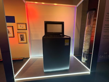 Lavadora negra con pantalla, caja de vidrio, plataforma iluminada. Tubo transparente con pelo. Marcos de fotos en pared. Cartel con texto