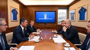 Seis hombres sentados en una mesa de madera. En el fondo, una pantalla muestra el logo de la Federación Italiana de Fútbol y una ventana con el Coliseo. Hay camisetas enmarcadas.